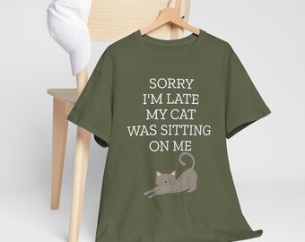 May include: Olive green t-shirt with the text "SORRY I'M LATE MY CAT WAS SITTING ON ME" in white. A gray cat illustration is below the text. A white baseball cap hangs on a wooden chair in the background.