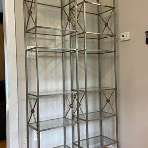 May include: Two tall, silver metal and glass display shelves. Each shelf has four glass shelves and decorative gold accents. The top of each shelf has a gold bird-like design. The shelves are standing on a wooden floor.