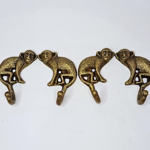May include: A set of four brass-colored metal wall hooks, each shaped like a seated monkey with a textured finish. The monkey's tail curves downward to form the hook portion, and each piece features a small circular opening for mounting purposes.