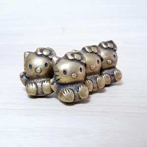 Könnte beinhalten: Vier kleine, messingfarbene Katzenfiguren mit Schleife, in einer Reihe angeordnet. Die Figuren haben einen Vintage-Look mit sichtbaren Details wie Gesicht, Pfoten und Schleife der Katze. Der Hintergrund ist eine hellfarbene Holzoberfläche.