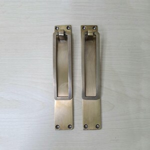 Könnte beinhalten: Zwei rechteckige Messing-Griffmuscheln mit gebürstetem Metall-Finish. Jeder Griff verfügt über einen klappbaren, vertieften Greifmechanismus und vier vorgebohrte Schraublöcher an der Ober- und Unterseite zur Montage auf einer flachen Oberfläche.