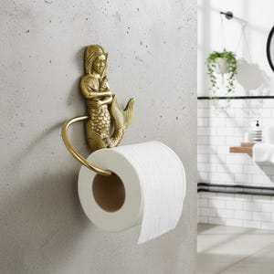 May include: A gold-toned metal toilet paper holder featuring a sculpted mermaid figure with a textured tail. The mermaid is positioned vertically, with a curved arm extending to support a standard roll of white toilet paper against a gray wall.