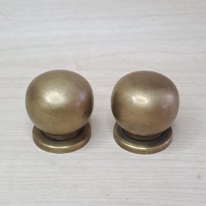 May include: Two spherical cabinet knobs made of brushed brass with a warm metallic finish. Each knob features a smooth, rounded top section that transitions into a circular base, providing a consistent look for furniture or door hardware.