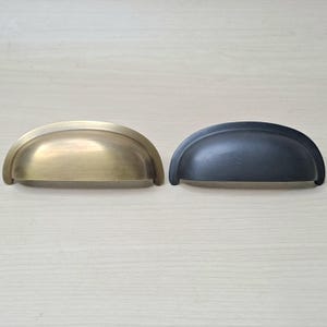 Op de afbeelding: Twee halvemaanvormige kastgrepen. Eén heeft een geborstelde messing afwerking, de andere is matzwart. Beide zijn gemaakt van metaal en hebben een gebogen ontwerp met een kleine lip aan elk uiteinde. Deze grepen zijn voor laden of kasten.