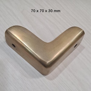 May include: An L-shaped furniture corner protector with a brushed brass finish. The metal component features two visible circular screw holes on its sides. The dimensions are clearly printed above the object as 70 x 70 x 30 mm.