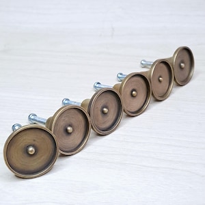 Könnte beinhalten: Sechs antike Messing-Schrankknöpfe mit kreisförmigem Design und einer zentralen Schraube. Die Knöpfe sind diagonal angeordnet und zeigen ihre gealterte, warmtonige Oberfläche. Ideal, um Möbeln einen Vintage-Touch zu verleihen.