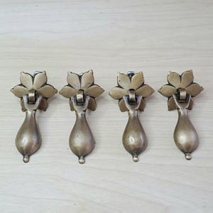 Könnte beinhalten: Vier antike Schubladengriffe aus Messing. Jeder Griff hat eine blütenförmige Rückplatte und einen tropfenförmigen Griff. Das Messing hat eine warme, gealterte Patina, die auf Vintage- oder handwerkliche Qualität hindeutet. Sie sind in einer Reihe auf einer hellen Holzoberfläche angeordnet.