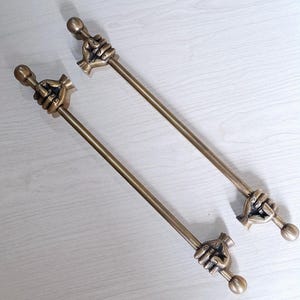 May include: Two long, slender, metallic bronze-toned rods, each featuring a sculpted hand at both ends. The hands are depicted in a closed-fist position, gripping the rod, with a small spherical finial extending from the wrist area of each hand.