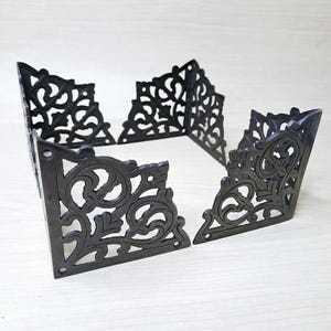 May include: Four black metal corner protectors featuring an intricate, openwork scroll pattern. Each piece is L-shaped with pre-drilled holes at the ends for attachment, showcasing a detailed, symmetrical design with curved lines and leaf-like motifs.