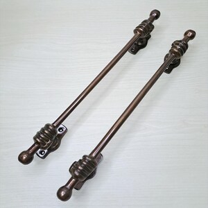 Könnte beinhalten: Zwei lange, bronzefarbene Metalltürgriffe mit einem glatten, zylindrischen Stangendesign. Jeder Griff verfügt über dekorative, gerippte, kugelförmige Endkappen und Montageplatten mit sichtbaren Schraublöchern an beiden Enden zur Befestigung.