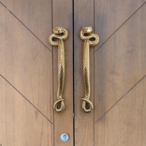 Puede incluir: Un par de manijas de puerta verticales con forma de serpientes enroscadas con un acabado dorado pulido. Cada manija presenta un patrón de escamas texturizado detallado, una cabeza curva en la parte superior y una cola en bucle en la parte inferior.
