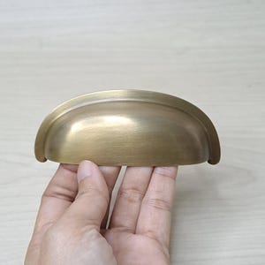 May include: A brass-colored, half-moon shaped drawer pull. The hardware has a brushed finish and is held by a hand. The drawer pull is designed to be mounted on a drawer or cabinet door. The hardware is made of metal.