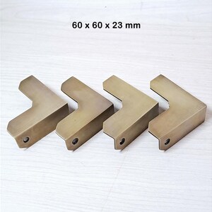 May include: Four L-shaped brass corner protectors with a brushed finish, each featuring a single pre-drilled screw hole on the side. The dimensions 60 x 60 x 23 mm are printed above the items, indicating the size of these metal hardware pieces.