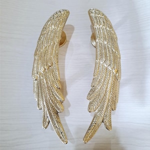 Puede incluir: Un par de manijas de puerta de metal color dorado con forma de alas de ave alargadas. Cada manija presenta grabados de plumas detallados y texturizados a lo largo de toda la longitud, con una base de montaje circular visible en la parte superior.