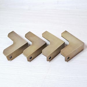 Puede incluir: Cuatro escuadras de esquina en forma de L de color latón. Cada escuadra tiene un pequeño agujero cerca de la esquina. Las escuadras están dispuestas en fila sobre una superficie de madera clara.