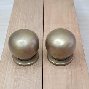 Pode incluir: Dois puxadores de armário esféricos de cor latão com base circular. Os puxadores estão colocados sobre uma superfície de madeira clara. Os puxadores têm um acabamento liso e fosco e são uniformemente espaçados.