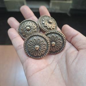 Puede incluir: Cuatro botones metálicos circulares con un patrón floral en relieve de doce pétalos que irradian desde un punto central. Los bordes de cada botón están decorados con un borde de cuentas y el metal tiene un acabado de bronce envejecido.
