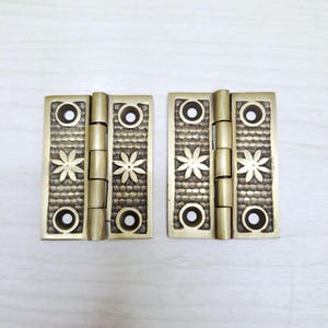 May include: Two antique brass hinges. Each hinge features a rectangular plate with a floral and circular design. The hinges are connected by cylindrical joints. The brass has a warm, aged patina.
