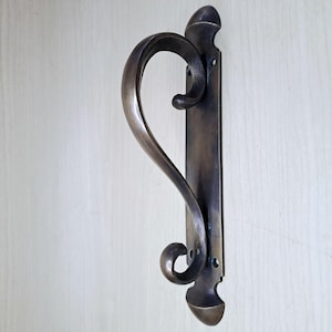 May include: Bronze-toned door pull with a curved handle. The handle has a graceful, flowing design, with a scroll-like shape. The backplate is rectangular with decorative details at the top and bottom. The hardware is mounted on a light-colored surface.