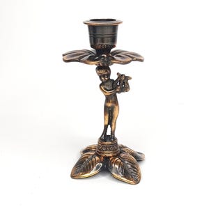 Könnte beinhalten: Ein bronzefarbener Kerzenhalter mit einem Cherub, der eine Leier spielt. Der Kerzenhalter hat einen blütenförmigen Sockel mit Blattdetails und eine Tasse für die Kerze. Der Cherub steht auf einem dekorativen Sockel. Gesamthöhe ca. 15 cm.
