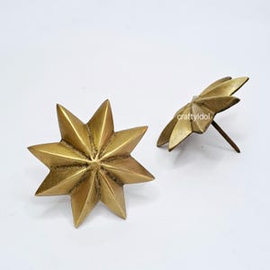 May include: Two brass-colored star-shaped decorative tacks. One tack is viewed from the front, displaying eight points. The other tack is viewed from the side, showing the pin. The text "craftyidol" is visible on the side of the second tack.