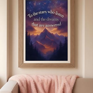 Puede incluir: Una impresión artística enmarcada con una escena de montañas y un cielo estrellado. La obra de arte presenta el texto "To the stars who listen and the dreams that are answered". El marco es de color marrón claro, y la impresión se muestra sobre una manta rosa.