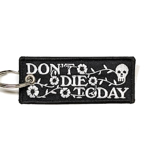 Könnte beinhalten: Schwarzer Schlüsselanhänger mit weißer Stickerei, die "Don't Die Today" mit einem Totenkopf und einem floralen Design zeigt.