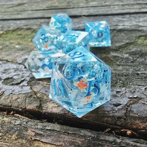 May include: A set of seven clear, blue-tinted polyhedral dice with embedded orange fish and blue numbers. The dice are arranged on a weathered wooden surface, showcasing their aquatic theme. Ideal for tabletop gaming.