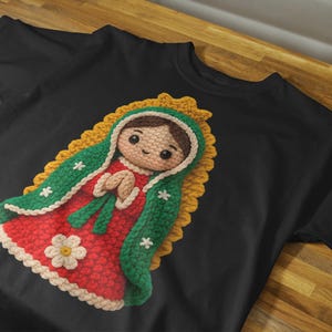 May include: Black t-shirt with a crochet design of a religious figure. The figure is wearing a red dress, green shawl, and a yellow halo. The design includes white flower and star accents.