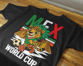 May include: Black t-shirt featuring a cartoon jaguar wearing a green jersey and a feathered headdress, kicking a soccer ball. The shirt has the word "MEX" in green, white, and red, and the words "WORLD CUP".