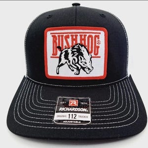May include: Black trucker hat with a white mesh back and a flat brim. The front features a rectangular patch with the words "BUSH HOG" in red and a black and white illustration of a wild boar. The hat has a Richardson tag.