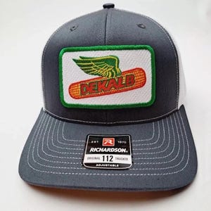 Peut inclure: Casquette de camionneur grise et blanche avec une visière incurvée. L'avant présente un écusson rectangulaire avec le mot "DEKALB" et un motif de maïs ailé. Le chapeau comprend une étiquette Richardson 112 Trucker.