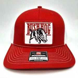 May include: Red and white trucker hat with a square patch featuring the text "RUSH HOG" above a black and white illustration of a hog. The hat has a curved brim and a mesh back. The tag reads "RICHARDSON ORIGINAL 112 TRUCKER ADJUSTABLE".