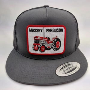Può includere: Cappellino da camionista grigio con visiera piatta e retro in rete. La parte anteriore presenta una toppa rettangolare con la scritta "MASSEY FERGUSON" sopra un'illustrazione di un trattore rosso e bianco. Un'etichetta bianca con testo nero è sulla visiera.