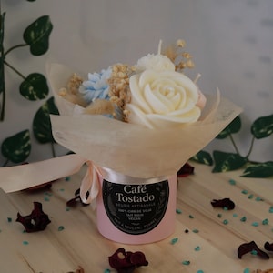 May include: A pink candle in a cylindrical container, wrapped with a bouquet of white, blue, and beige flowers. The candle is tied with a pink ribbon and has a label that reads "Café Tostado".