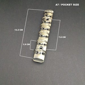 May include: A bronze-colored metal six-ring binder mechanism for A7 pocket size paper. The component features a total length of 13.3 cm, a ring diameter of 1.9 cm, and a distance of 7.6 cm between the two mounting holes for attachment.