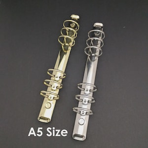May include: Two A5 size binder mechanisms, one gold-toned and one silver-toned, are shown against a dark background. Each binder has multiple metal rings for holding paper. The text "A5 Size" is printed in white.