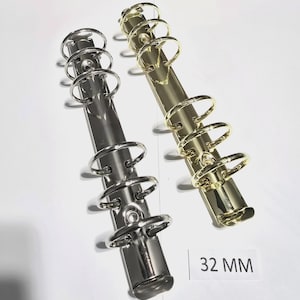 Puede incluir: Dos anillas metálicas, una plateada y otra dorada, se muestran sobre un fondo blanco. Las anillas miden 32 mm. Las anillas están diseñadas para su uso en cuadernos y planificadores.