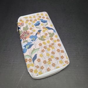 May include: A floral print wallet with a zipper closure. The wallet features a light pink background with a pattern of blue, yellow, and brown flowers and green leaves. The zipper is silver.
