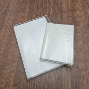 May include: Two clear plastic document holders, one larger and one smaller, are stacked on a wooden surface. The holders have a transparent design, allowing the white paper inside to be visible. The holders are designed to protect and organize documents.