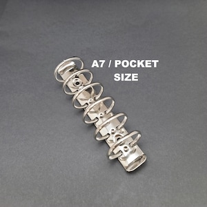 May include: A silver-colored A7 pocket size binder mechanism. The binder has six circular rings and is made of metal. The text "A7 / POCKET SIZE" is printed in white.