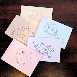 Puede incluir: Una colección de cinco pequeñas ilustraciones dibujadas a mano sobre papel blanco y rosa. Los dibujos presentan bocetos caprichosos de gatos, zorros y otras figuras de dibujos animados. La obra de arte está sobre una superficie de madera marrón oscuro.