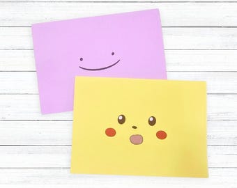 May include: Two rectangular cards with cartoon faces. One is lavender with a smiling face. The other is yellow with a surprised face, red cheeks, and a small mouth. The cards are on a white wooden surface.