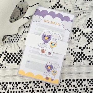 May include: A notepad with a white background and a decorative band featuring cartoon characters. The notepad has a purple and yellow cloud design with the text "GET EM ALL". The notepad is designed for taking notes.