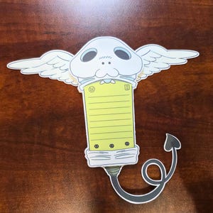 May include: A whimsical die-cut sticker featuring a cartoonish design. The sticker has a light yellow notepad with a smiling face and a small illustration. It has a white head with wings, and a black tail with a heart-shaped tip.