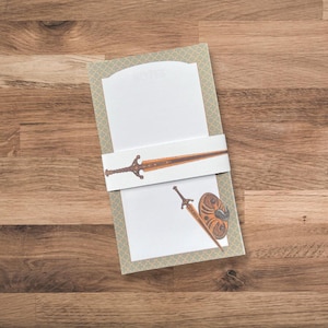 May include: A notepad with a decorative border and a white band featuring a sword illustration. The notepad has a sword and shield illustration on the bottom page. The word "NOTES" is at the top.