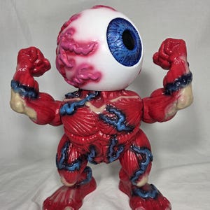 Puede incluir: Una figura &uacute;nica de un ojo musculoso con detalles de m&uacute;sculos rojos, venas azules y un gran globo ocular blanco con iris azul. La figura posa con los brazos y pies flexionados, mostrando su dise&ntilde;o anat&oacute;mico.