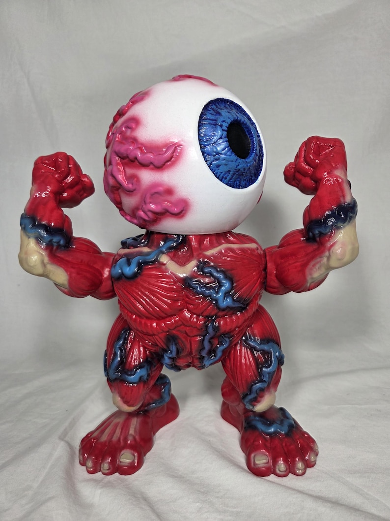 Puede incluir: Una figura &uacute;nica de un ojo musculoso con detalles de m&uacute;sculos rojos, venas azules y un gran globo ocular blanco con iris azul. La figura posa con los brazos y pies flexionados, mostrando su dise&ntilde;o anat&oacute;mico.