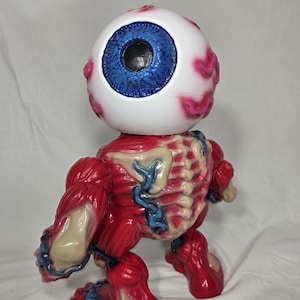 Puede incluir: Una figura &uacute;nica de un globo ocular estilizado con iris azul y escler&oacute;tica blanca, detallado con venas rosas. El cuerpo es rojo, representando m&uacute;sculos y estructura &oacute;sea, con venas azules y detalles &oacute;seos beige. La figura est&aacute; de pie.