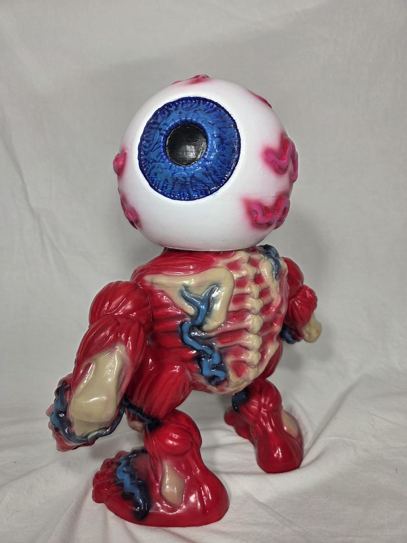 Puede incluir: Una figura &uacute;nica de un globo ocular estilizado con iris azul y escler&oacute;tica blanca, detallado con venas rosas. El cuerpo es rojo, representando m&uacute;sculos y estructura &oacute;sea, con venas azules y detalles &oacute;seos beige. La figura est&aacute; de pie.